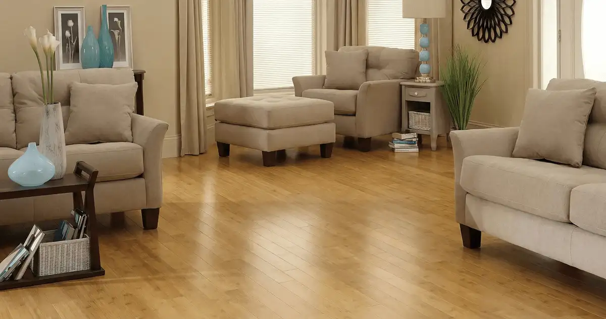 A living room with professionally installed bamboo flooring, beige sofas, a matching ottoman, a side table with a blue lamp, vases, framed pictures, and large windows with blinds and curtains in Honolulu, HI